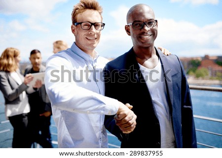 Black male colleague and white male colleague smiling at camera and shaking hands