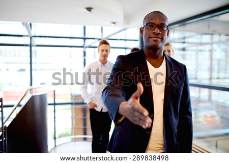Young black executive facing camera with hand stretched out and colleagues in the background.
