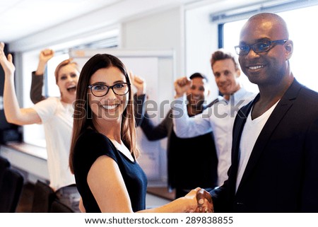 White female colleague and black male colleague shaking hands in front of coworkers flexing arms