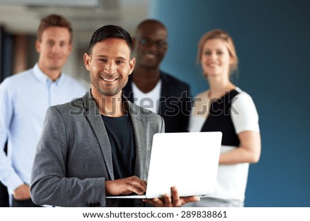 Young white executive holding laptop smiling at camera with colleagues in background