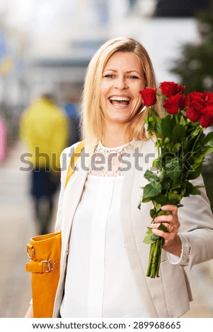 White woman posing on sidewalk grinning holding a bouquet of red roses