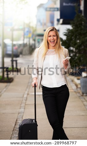 Professional white woman grinning holding her cellphone and posing on sidewalk while holding suitcase handle