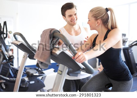 Handsome Young Fitness Trainer Explaining Something to a Young Woman While on Elliptical Bike Device Inside the Gym