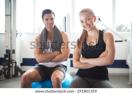Young Fit Couple Sitting on Exercise Balls Inside the Gym and Smiling at Camera with Arms Crossing Over their Stomach.