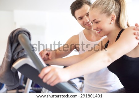 Sweet Athletic Handsome Boyfriend Assisting his Pretty Girlfriend Doing an Exercise on Elliptical Bike Inside the Gym