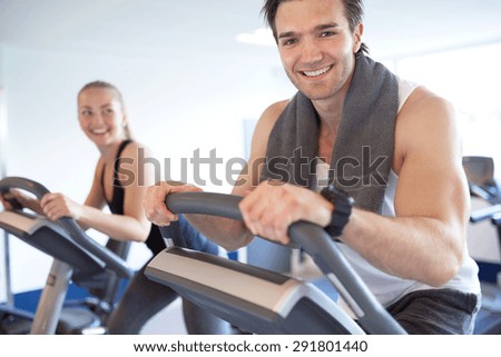 Handsome Muscular Young Guy Smiling at the Camera, While Exercising on Elliptical Bike, with his Partner Next to him Inside the Fitness Gym.