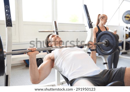 Muscular Man Lifting Barbell Weight, Doing Chest Press, While Woman Lifts Hand Weight in Background of Brightly Lit Gym
