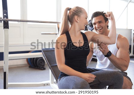 Male Trainer Testing Size of Blond Woman Bicep Muscle, Playful Couple Checking Muscles and Smiling at Each Other in Brightly Lit Gym