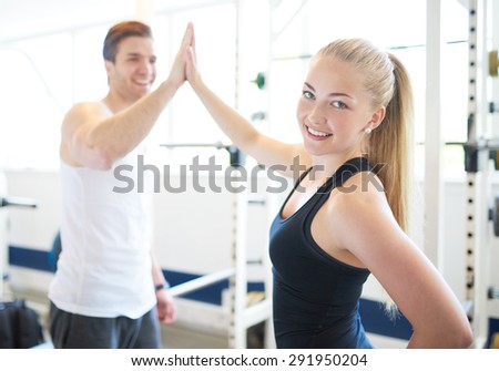 Blond Woman Looking Over Shoulder at Camera and High Fiving Male Trainer in Gym, Celebrating Accomplishment of Fitness Goal