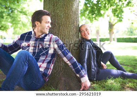 Sweet Young Lovers Holding Their Hands and Smiling to Each Other While Relaxing Under the Shade of a Tree at the Park.