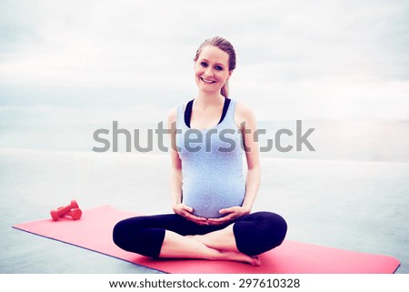 Happy woman keeping fit during her pregnancy sitting cross legged on an exercise mat cradling her swollen belly with dumbbells alongside her as she smiles at the camera
