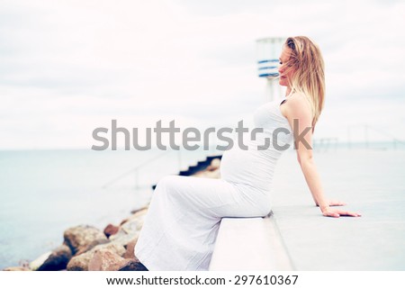 Pregnant young woman relaxing at the seaside sitting on a promenade overlooking the ocean in a white maternity dress, side view emphasising her swollen belly