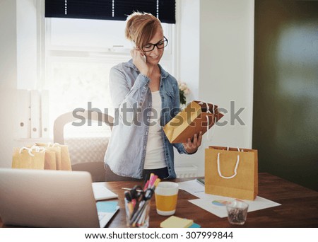 Motivated businesswoman placing orders on the phone for the packaging for her new online web based store as she holds a brown paper gift bag in her hand