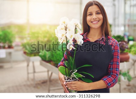 Smiling happy young florist in her nursery standing holding a potted white Phalaenopsis orchid plant in her hands as she tends to the houseplants in the greenhouse