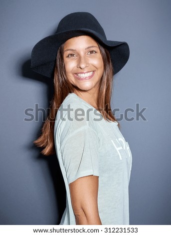 Cute young woman with a big beaming smile wearing a trendy felt hat standing grinning at the camera over grey in a fun humorous portrait