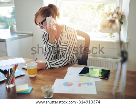 Young manageress working at her desk in the office taking a call on her mobile phone while writing notes on a notepad, charts and graphs in the foreground