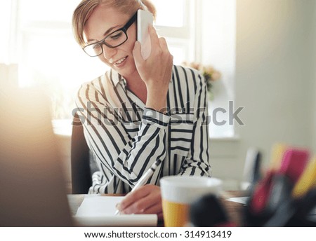 Successful self-employed young woman working from a home office taking an order over the mobile phone from products on her website and writing it down on a notepad