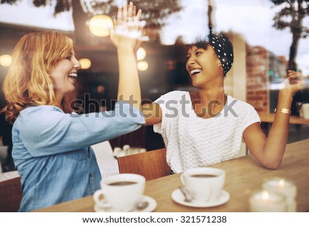 Happy exuberant young girl friends giving a high five slapping each others hand in congratulations as they sit together in a cafeteria enjoying a cup of hot coffee, multi ethnic viewed through glass