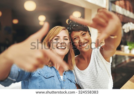 Vivacious young women framing their faces with a finger frame in a fun creative gesture as they sit in a cafeteria together, young multiethnic couple