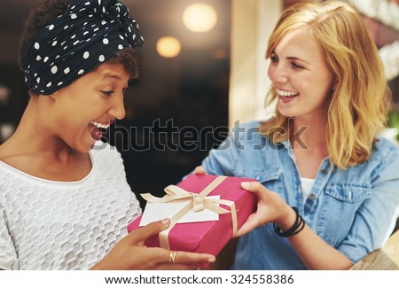 Pretty young blond woman giving an attractive young African American friend a surprise gift gift-wrapped with red paper, a bow and card