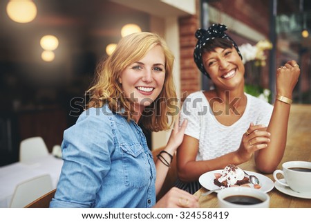 Two attractive young multiethnic female friends enjoying coffee and cake together in a cafeteria smiling happily at the camera as they sit together at a table