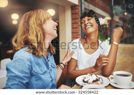 Two multiethnic young female friends enjoying coffee together in a restaurant laughing and joking while touching to display affection