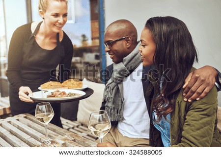 Waitress serving food to an affectionate African American couple sitting arm in arm at a restaurant table