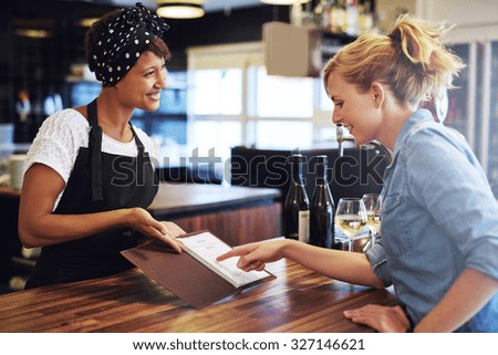 Female customer choosing wine from a wine list being presented to her by a charming young African American bartender in a bar conceptual of employment, small business ownership or an entrepreneur