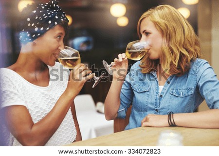 Two attractive young women meeting up in a pub for a glass of white wine sitting at a counter smiling at each other as they sip their glasses