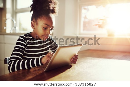 Young black girl with a fun afro hairstyle sitting at a table at home browsing the internet on a tablet computer with bright sun flare through the window alongside her