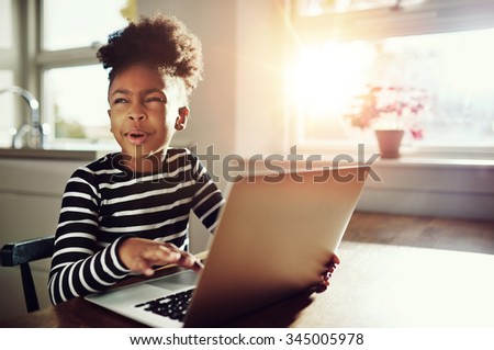 Fun little black girl with her frizzy afro hair up on top of her head sitting at a table at home working on a laptop computer screwing her eyes up in thought as she tries to think of an answer