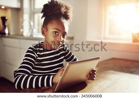 Thoughtful young black girl sitting watching the camera with a pensive expression as she browses the internet on a tablet computer at home