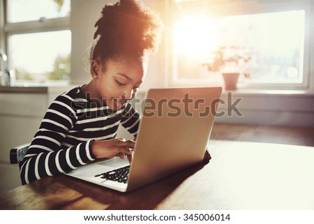 Ethnic girl with a cute afro hairstyle sitting at home using a laptop computer to do her class work and browse the internet, bright sun glow behind