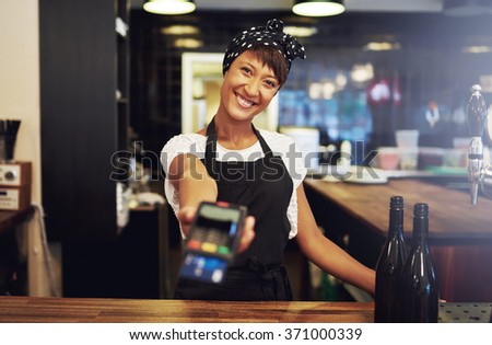 Charismatic young female small business owner requesting payment form a client holding out a handheld banking machine to process their credit card, focus to her face and warm friendly smile
