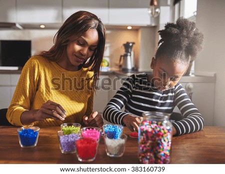 Child and parent sitting and creating color beaded crafts on wooden table in kitchen with various jars in front of them