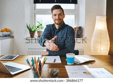 Smiling businessman leaning across his desk offering his hand in greeting, to close a deal, in partnership or congratulations