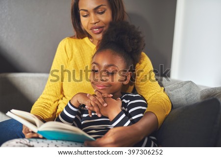 Black mom and daughter reading a book sitting on sofa smiling
