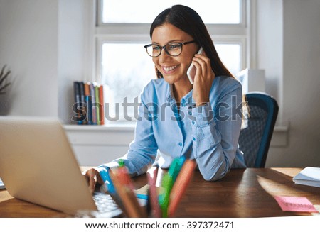 Single happy female business owner with smile and eyeglasses on phone and working on laptop computer at desk with bright window in background
