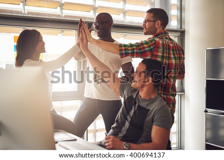 Group of four diverse men and women in casual clothing celebrating business accomplishments in office near desk and bright window