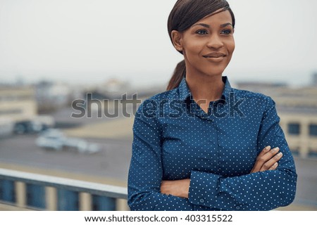 Confident friendly black business woman standing with folded arms on the rooftop of an urban commercial building smiling as she looks to the side of the camera