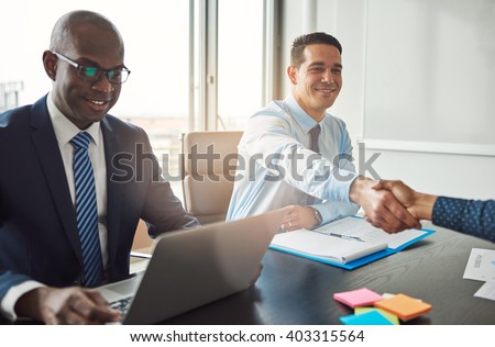 Smiling young Hispanic business man and woman shaking hands across a table in the office watched by a smiling African American manager