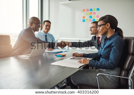 Two young multiracial business teams reaching an agreement in negotiations stretch across the table in the conference room to shake hands