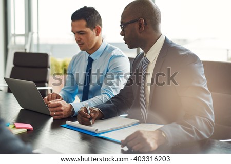 Two experienced business executives in a meeting seated at a table discussing paperwork and information on a laptop computer, one Hispanic, one African American