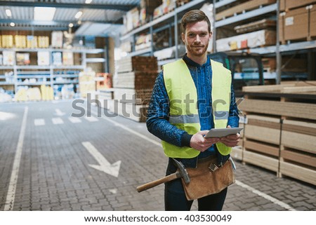 Handsome young handyman or warehouse supervisor standing amongst the building supplies with a tablet in his hand smiling at the camera