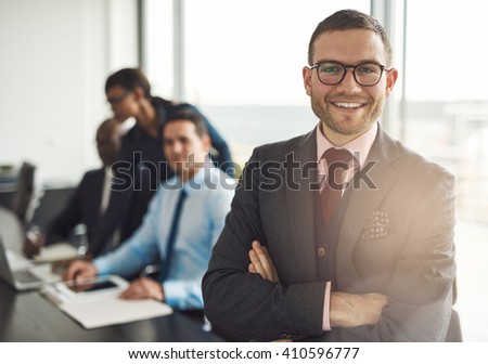 Confident smiling business executive with folded arms near conference table with three co-workers discussing something in large bright office room