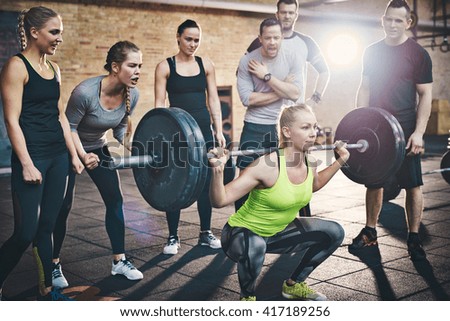 Fit young woman lifting barbells looking focused, working out in a gym with other people cheering her on
