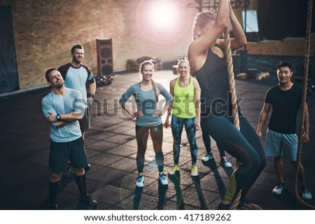 Fit young women climbing a rope in a gym with people on the floor watching