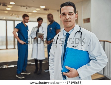 Thoughtful young Hispanic doctor in a hospital standing looking at the camera with a file under his arm as his multiracial team have a discussion in the background