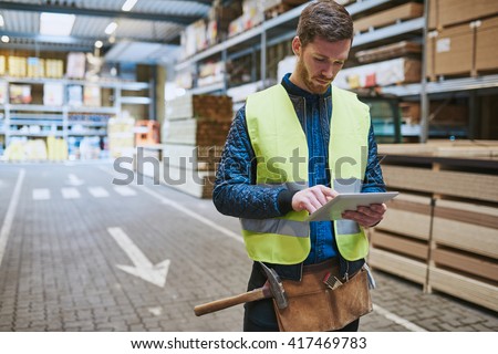 Young warehouse worker dealing in building supplies standing in a drive through consulting a handheld tablet computer