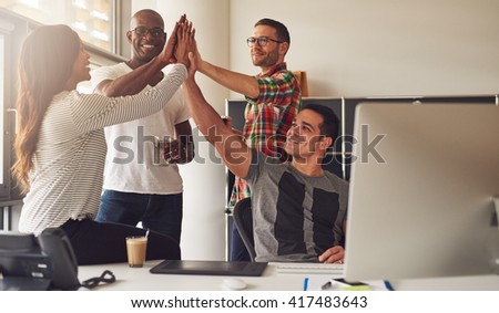 Group of four diverse men and women in casual clothing celebrating an important milestone while relaxing in office near large bright window